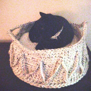 May include: A cream-colored, hand-crocheted pet bed with a textured cable knit design. The bed has a round shape with two handles and is filled with a soft, light-colored cushion. A black cat is curled up inside the bed.