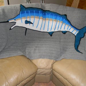 Könnte beinhalten: Eine gehäkelte Marlin-Decke in Blau-, Weiß- und Beigetönen, vor einem grauen Hintergrund. Die Decke zeigt eine detaillierte Darstellung eines Marlin-Fisches mit seinem charakteristischen langen Schnabel und der Rückenflosse. Die Decke liegt auf einem beigefarbenen Ledersofa.