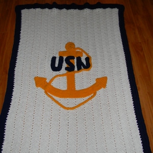 May include: A white crochet blanket with a navy blue scalloped border. The blanket features a large anchor design in orange and navy blue, with the letters "USN" above the anchor. The blanket is likely handmade.
