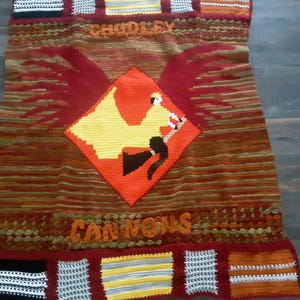 May include: A handmade, crocheted blanket featuring a central diamond design with a figure riding a broom, wings, and the words "CHUDLEY CANNONS". The blanket has a color scheme of red, orange, yellow, and brown.