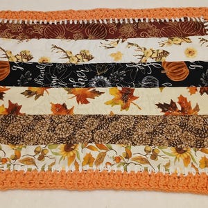 Könnte beinhalten: Eine rechteckige, gesteppte Stoffmatte mit horizontalen Streifen in herbstlichen Mustern, darunter Kürbisse, Sonnenblumen, Tannenzapfen und Herbstblätter. Die Matte ist mit einem dicken, strukturierten, orangefarbenen Häkelrand versehen.