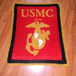 May include: A crocheted blanket featuring the letters "USMC" in gold, above a gold emblem with a white globe and eagle on a red background. The blanket has a black border with gold trim.