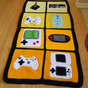 Può includere: Coperta all'uncinetto fatta a mano con un motivo a griglia di quadrati gialli e crema. Ogni quadrato presenta un diverso design di controller per videogiochi in bianco, nero e verde. Bordo nero.