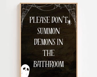 May include: A framed sign with a black background and white text that reads "PLEASE DON'T SUMMON DEMONS IN THE BATHROOM." A white spider web and spider are at the top, and a cartoon ghost is in the lower left corner. The frame is light brown.
