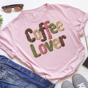 May include: A light pink t-shirt with the words "Coffee Lover" in a textured, multi-colored design. The text features brown, pink, green, and beige colors. The shirt is folded, with a pair of sunglasses and denim jacket visible.