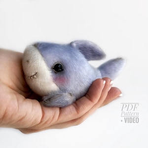 May include: A handmade plush shark toy in shades of blue and white, with a stitched mouth and black eyes. The shark has a blush of pink on its cheeks and is held in a person's hand. The toy is soft and cuddly.