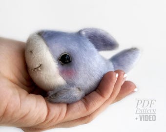 May include: A handmade plush shark toy in shades of blue and white, with a stitched mouth and black eyes. The shark has a blush of pink on its cheeks and is held in a person's hand. The toy is soft and cuddly.
