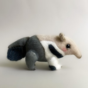 May include: A plush anteater toy in shades of gray, white, and beige. The toy has a long snout, small ears, and a fluffy tail. The anteater's cheeks are lightly blushed, and it has large, black eyes.