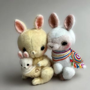 May include: Two plush bunny dolls. One is yellow with a smaller white bunny nestled in its arms. The other is white with a colourful embroidered shawl. Both have large black eyes and pink-lined ears.