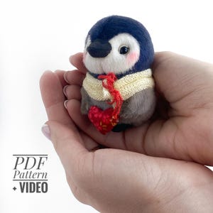 May include: A plush penguin with a blue head, white face, and gray body, wearing a cream-colored scarf and holding a red heart. The penguin has rosy cheeks and a black beak. The image includes the text "PDF Pattern + VIDEO".
