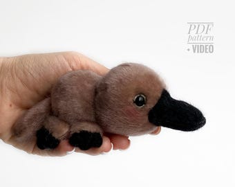 May include: A plush platypus toy with a brown body, black beak, and black feet. The toy has small, black eyes and a hint of pink on its cheeks. The image includes the text "PDF pattern + VIDEO".