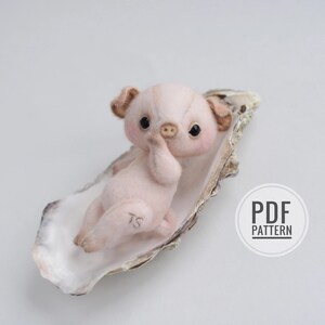 May include: A handmade, light pink piglet doll with dark eyes and a small snout, resting in a large oyster shell. The piglet has small, tan ears and a tiny hand raised to its face. The shell is a light beige color. The image includes a "PDF PATTERN" logo.