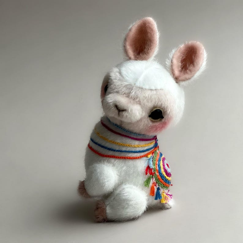 May include: A white plush llama toy with pink inner ears and rosy cheeks. It is adorned with a colourful, embroidered scarf featuring rainbow stripes and tassels. The llama is seated, with its legs tucked in, and has large, dark eyes.