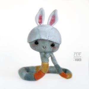 May include: A soft toy snake wearing a bunny hat. The snake is mainly grey with a segmented body in shades of blue, orange, and yellow. The bunny hat is white with pink inner ears. The toy has a smiling face and large black eyes. Text on the right says "PDF pattern + VIDEO".