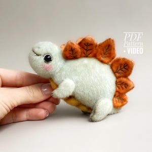 以下が含まれることがあります： パステルカラーの柔らかいステゴサウルスのぬいぐるみ。恐竜は、薄緑色の体、オレンジ色のプレート、黄色の腹を持っています。おもちゃは、バラ色の頬と小さな笑顔で甘い表情をしています。画像には「PDF Pattern + VIDEO」というテキストも含まれています。