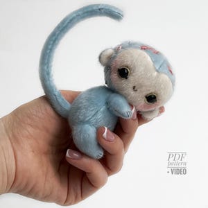Può includere: Una piccola scimmia di peluche blu con una lunga coda curva. La scimmia ha un viso bianco, grandi occhi neri e guance rosa. È tenuta in una mano. Il testo "PDF pattern + VIDEO" è visibile.