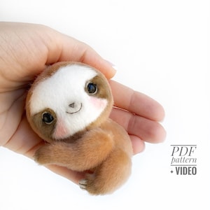 May include: A handmade plush sloth toy with a white face, brown eyes, and rosy cheeks. The sloth has a light brown body and is held in a hand. The image includes the text "PDF pattern + VIDEO".