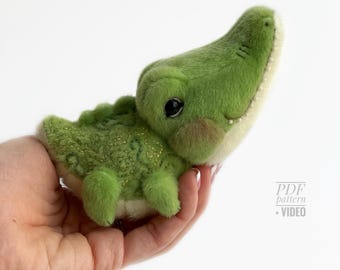 May include: A plush green alligator toy with a light green belly and a textured back. The alligator has black eyes and a smiling expression. The toy is held in a hand against a white background. The text "PDF pattern + VIDEO" is in the bottom right corner.
