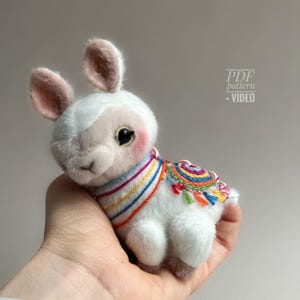 May include: A white plush llama toy with pink ears and rosy cheeks, wearing a colourful embroidered blanket and a rainbow-striped collar. The text "PDF pattern + VIDEO" is visible on the right.