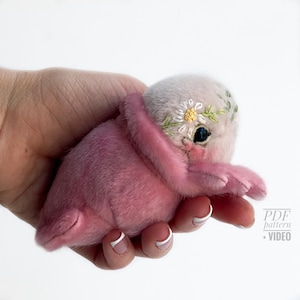 May include: A handmade pink plush creature with a daisy crown and embroidered details. The plush toy is held in a hand, showcasing its soft texture and intricate design. The creature has a light pink body and a grey head.