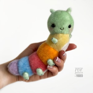 May include: A plush, colorful caterpillar toy with a smiling face. The caterpillar's body transitions through shades of green, yellow, orange, red, blue, and purple. It has small, green feet and antennae. The toy is held in a hand.