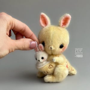 Puede incluir: Un peluche de conejo amarillo con un conejito blanco más pequeño metido en su bolsa. El conejo más grande tiene orejas forradas de rosa y mejillas rosadas. La imagen incluye el texto "PDF pattern + VIDEO".
