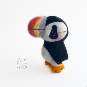 Puede incluir: Un peluche de frailecillo hecho a mano con un pico grande y colorido con rayas rojas, amarillas y azules. El ave tiene plumas negras y blancas, patas naranjas y un rubor rosado en las mejillas. El texto "PDF Pattern + VIDEO" está en la esquina inferior izquierda.