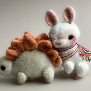 May include: Two plush toys: a light green dinosaur with orange spines and a white llama with a colourful embroidered shawl. The dinosaur has a soft, textured appearance, while the llama has long ears and a sweet expression. The toys are set against a plain background.
