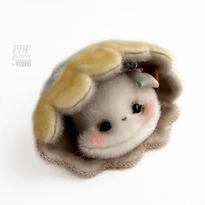 May include: A plush, light gray creature with rosy cheeks and black eyes, nestled under a yellow and blue shell. The shell is edged with a beaded trim. The image includes the text "PDF pattern + VIDEO".