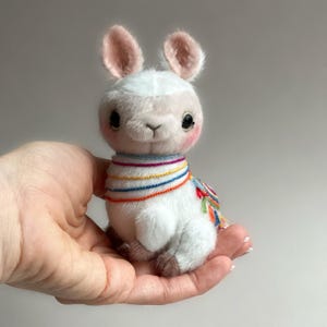 May include: A white llama plush toy with pink ears and rosy cheeks, wearing a colourful embroidered scarf and a decorative piece on its back. The llama is held in a hand.
