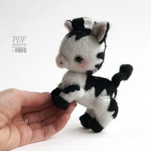 Puede incluir: Un juguete de peluche de vaca hecho a mano en blanco y negro, sostenido por una mano. La vaca tiene un diseño lindo con mejillas rosadas y una pequeña cola. La imagen también incluye el texto "PDF pattern + VIDEO".