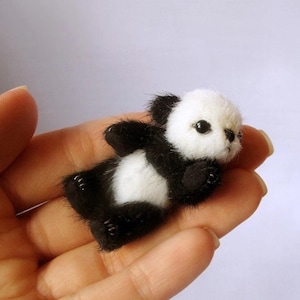 Op de afbeelding: Een miniatuur panda beer knuffel, met een zwart-wit kleurenschema. De panda heeft een pluizige witte kop, zwarte oren en zwarte ledematen. Het speelgoed ligt, met zijn kleine zwarte ogen zichtbaar.