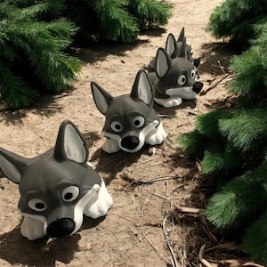 Puede incluir: Tres esculturas de cabezas de lobo grises y blancas dispuestas en fila sobre una superficie de tierra, con follaje verde de fondo. Las esculturas de lobos tienen ojos grandes y expresivos y están tumbadas. Una decoración de jardín caprichosa.