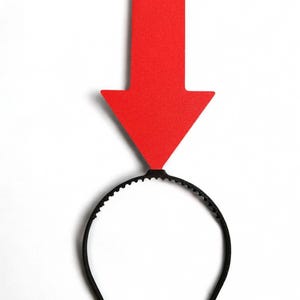 May include: A red arrow-shaped headpiece attached to a black headband. The arrow is a solid, vibrant red, pointing downwards. The headband is a simple, curved design, suitable for costume or novelty wear.