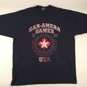 May include: A navy blue t-shirt with a red and white maple leaf design. The text "CAN-AMERA GAMES 19 94" is printed above the maple leaf. The text "SAGNAIN, THOMAS AND JAMES TOWNSHIPS, MICHIGAN, CAMBRIDGE ONTARIO, USA" is printed below the maple leaf.