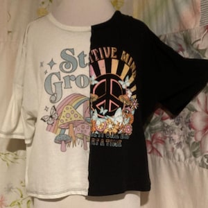 Puede incluir: Una camiseta blanca y negra con un diseño dividido. El lado blanco presenta un arcoíris, hongos, mariposas y el texto "Stay Gro". El lado negro presenta un signo de paz, flores, mariposas y el texto "Positive Mind Take It One Day At A Time".