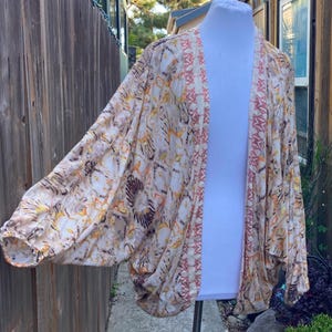 Può includere: Un cardigan stile kimono fantasia nei toni del beige, marrone e arancione. Il design aperto sul davanti presenta un motivo geometrico ripetuto ed è esposto su un manichino bianco. Le maniche sono ampie e fluide, offrendo una vestibilità rilassata.