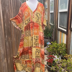 Puede incluir: Vestido hasta la rodilla con cuello en V y mangas cortas. El vestido presenta un diseño de patchwork en tonos naranja, amarillo y marrón, con estampados florales y geométricos. El vestido se exhibe en un maniquí al aire libre.