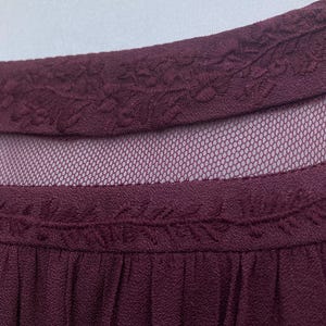 Puede incluir: Primer plano de un top burdeos con un escote bordado con motivos florales. La prenda presenta un panel de malla transparente y un dise&ntilde;o plisado, ofreciendo un aspecto texturizado y elegante.