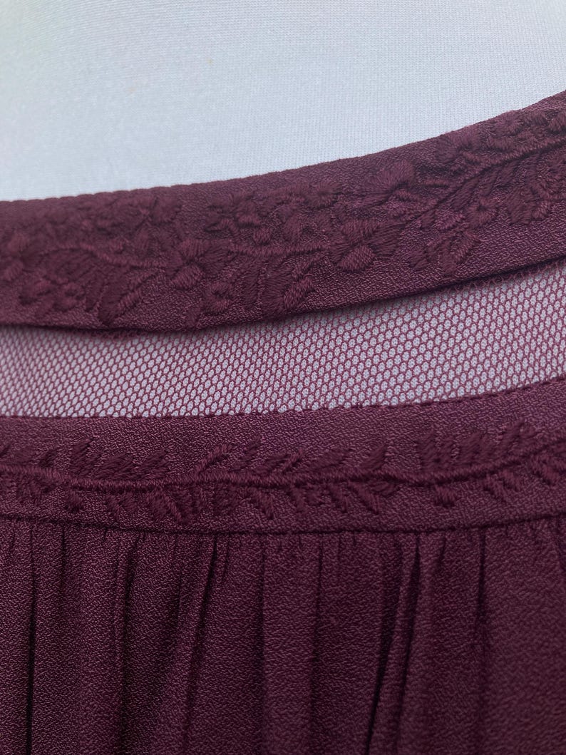 Puede incluir: Primer plano de un top burdeos con un escote bordado con motivos florales. La prenda presenta un panel de malla transparente y un dise&ntilde;o plisado, ofreciendo un aspecto texturizado y elegante.