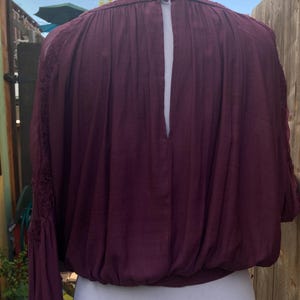 Puede incluir: Una blusa burdeos con mangas largas y cintura fruncida. La parte trasera de la blusa tiene una abertura de ojo de cerradura. Las mangas tienen detalles de encaje cerca de los hombros. La blusa se exhibe en un maniqu&iacute; blanco.