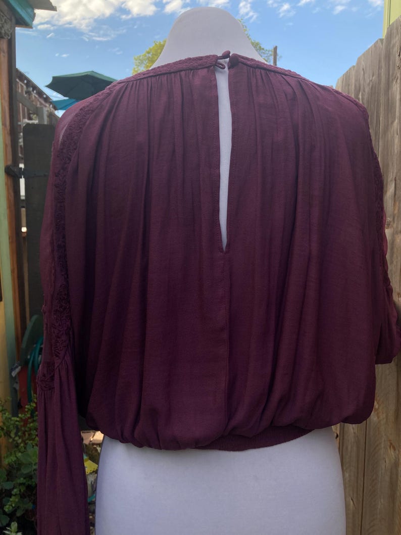 Puede incluir: Una blusa burdeos con mangas largas y cintura fruncida. La parte trasera de la blusa tiene una abertura de ojo de cerradura. Las mangas tienen detalles de encaje cerca de los hombros. La blusa se exhibe en un maniqu&iacute; blanco.