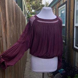 Puede incluir: Una blusa color ciruela con mangas largas y abullonadas y un escote transparente. La parte superior presenta intrincados bordados en las mangas y una cintura fruncida. La blusa se exhibe en un maniqu&iacute; blanco.