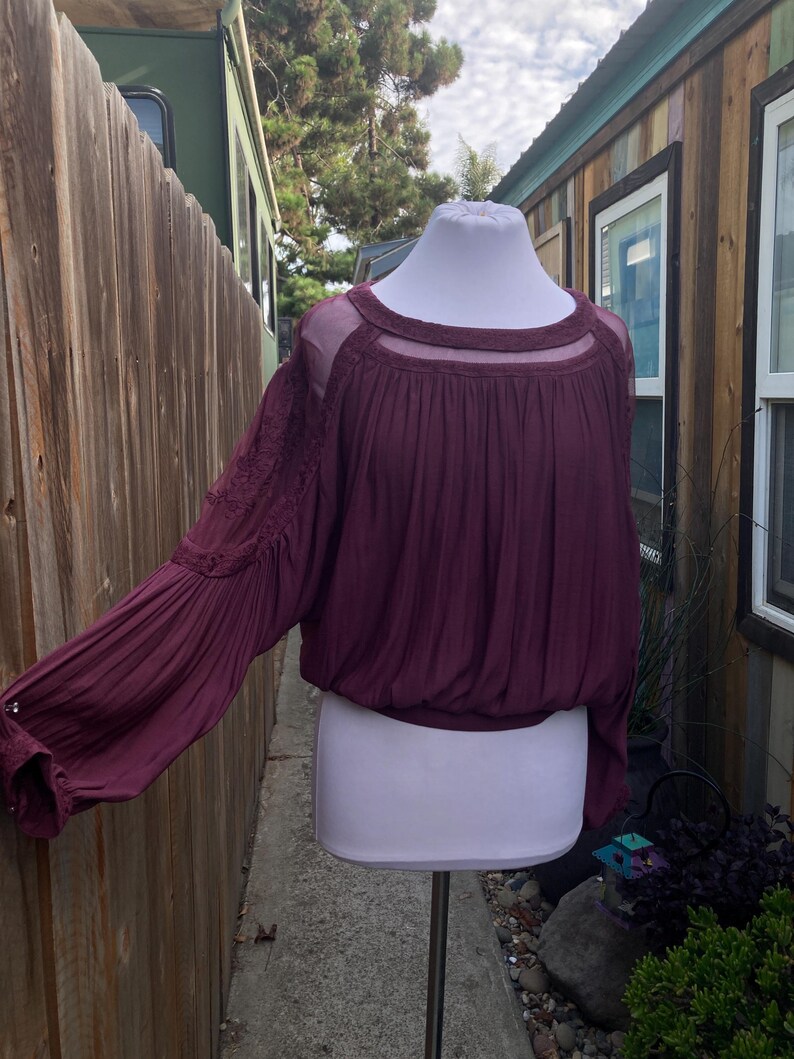 Puede incluir: Una blusa color ciruela con mangas largas y abullonadas y un escote transparente. La parte superior presenta intrincados bordados en las mangas y una cintura fruncida. La blusa se exhibe en un maniqu&iacute; blanco.