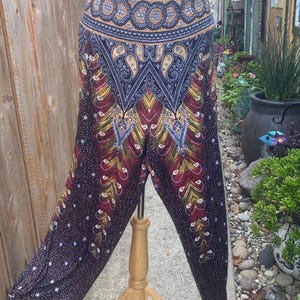 May include: A pair of patterned harem pants with a dark background and a colorful design. The pants feature a paisley and feather-like pattern in shades of red, yellow, blue, and gold. The pants are displayed on a wooden stand.