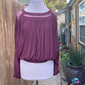 Puede incluir: Una blusa color ciruela con mangas largas y cuello redondo. La parte superior presenta un canes&uacute; transparente y detalles de encaje en las mangas. La blusa est&aacute; fruncida en la cintura y tiene un ajuste relajado, adecuado para ropa casual.