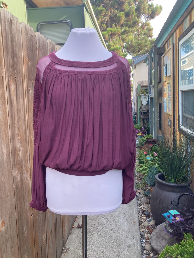 Puede incluir: Una blusa color ciruela con mangas largas y cuello redondo. La parte superior presenta un canes&uacute; transparente y detalles de encaje en las mangas. La blusa est&aacute; fruncida en la cintura y tiene un ajuste relajado, adecuado para ropa casual.