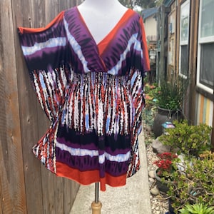 May include: A colorful, patterned dress with a V-neck and elastic waist is displayed on a mannequin. The dress features a mix of red, purple, white, and blue vertical stripes and a solid orange trim. The sleeves are wide and flowing.