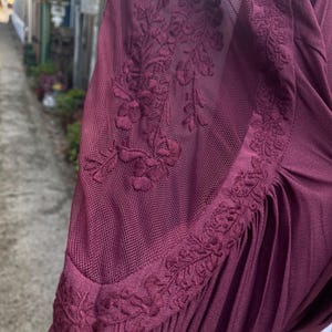 Puede incluir: Prenda de color burdeos con una manga de malla transparente. La manga presenta un intrincado bordado floral en el hombro y la parte superior del brazo. La tela parece suave y fluida, con sutiles pliegues.