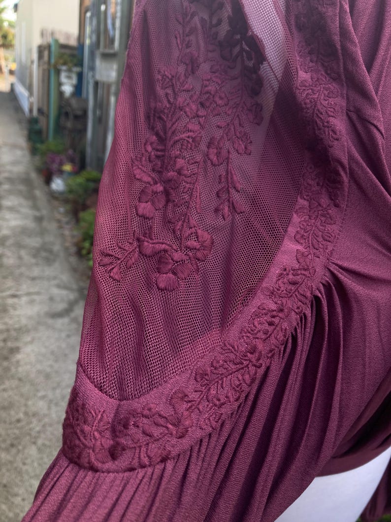 Puede incluir: Prenda de color burdeos con una manga de malla transparente. La manga presenta un intrincado bordado floral en el hombro y la parte superior del brazo. La tela parece suave y fluida, con sutiles pliegues.